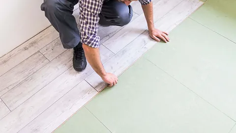 En mann legger lyst laminatgulv over grønt underlag som en del av oppussing av rommet.